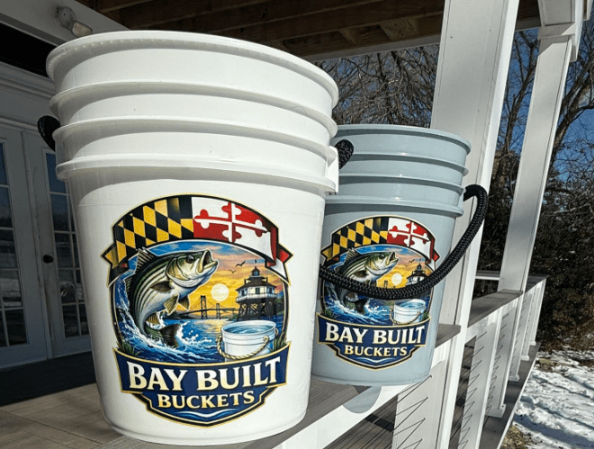 A stack of white and light blue buckets with the logo 'Bay Built Buckets,' featuring a fish and a scenic background with a lighthouse.